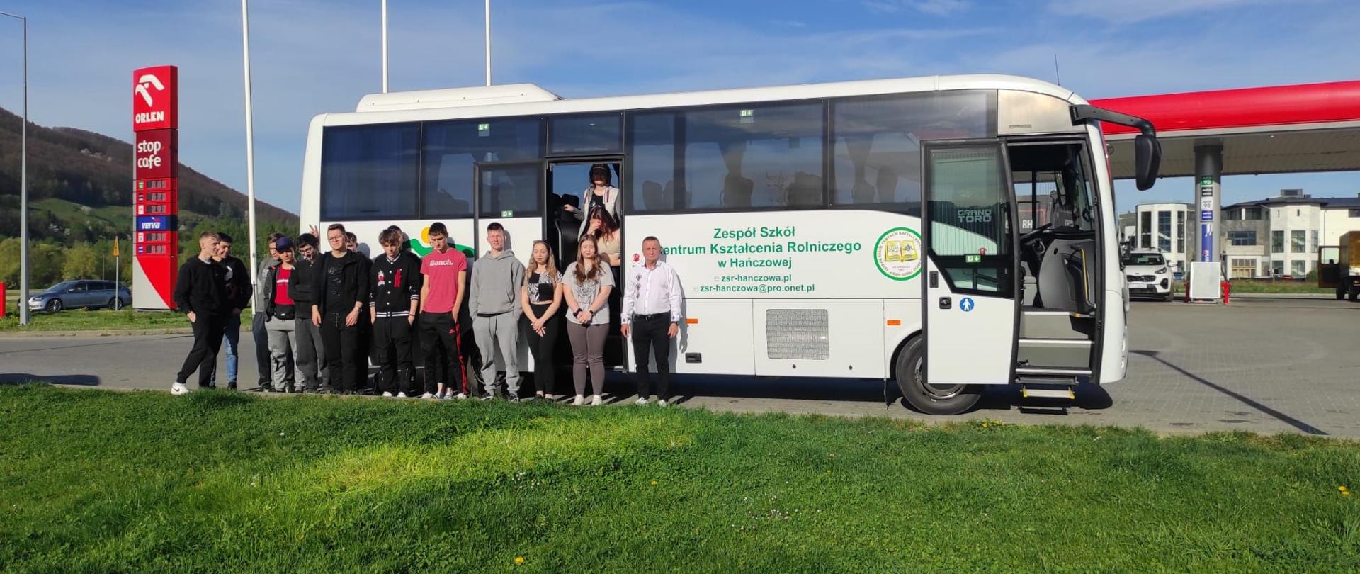 Uczniowie i opiekun stoją przed białym autokarem z napisem „Zespół Szkół Centrum Kształcenia Rolniczego w Hańczowej”. W tle widoczna jest stacja paliw Orlen oraz zielone wzgórza.