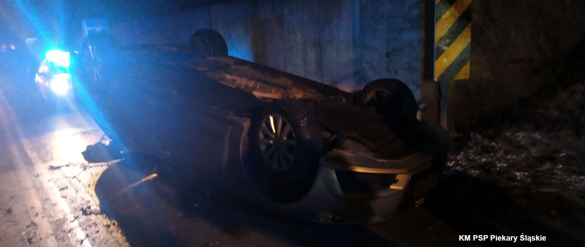 Zdjęcie przedstawia samochód osobowy marki Audi leżący pod wiaduktem, na drodze, na dachu. Z tyłu widać światła ostrzegawcze policyjnego radiowozu.