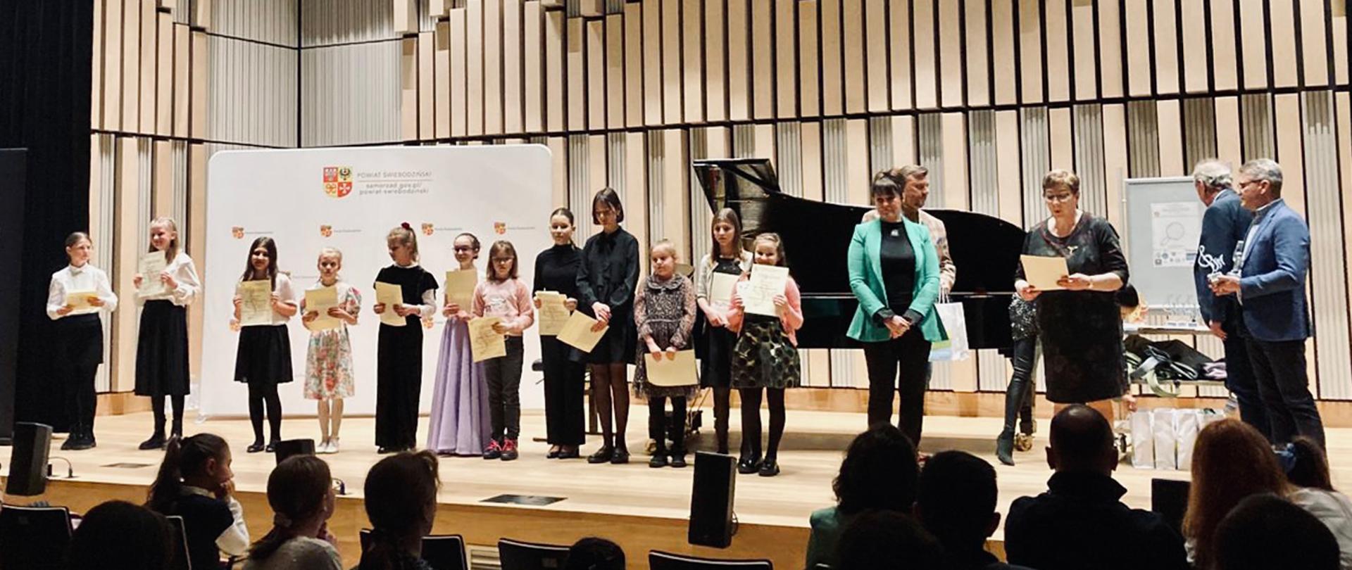 Na zdjęciu grupa kilkunastu osób - dorośli i dzieci stoją na scenie, większość z nich trzyma w rękach dyplomy. Za nimi stoi fortepian.