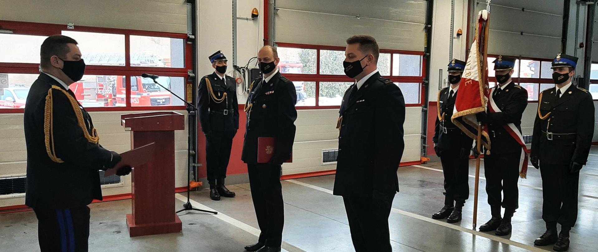 Zdjęcie ilustruje uroczystość przekazania obowiązków komendanta powiatowego PSP w Żywcu. W garażu Komendy Powiatowej w Żywcu z lewej strony stoi w mundurze galowym i maseczce ochronnej Śląski Komendant Wojewódzki PSP nadbryg. Jacek Kleszczewski, który w ręce trzyma akt powołania. Po prawej stronie ubrani w mundury galowe i maseczki ochronne stoją: odchodzący na zaopatrzenie emerytalne st. bryg. Krzysztof Grygiel i nowo powołany Komendant Powiatowy PSP w Żywcu, mł. bryg. Łukasz Węgliński. W głębi stoi poczet sztandarowy.