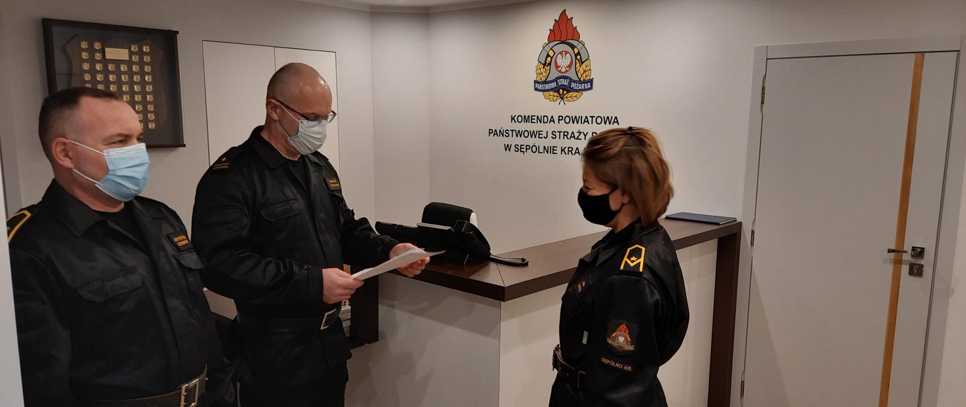zdjęcie przedstawia z lewej strony stoją bokiem mł. asp. Krystian Szwarc obok niego mł. bryg. Dariusz Chylewski Zastępca Komendanta Powiatowego PSP, który odczytuje list gratulacyjny z okazji 20 lecia pracy i służby Anny Siekierki. Naprzeciwko Z-cy Komendanta bokiem stoi mł. asp. Anna Siekierka. Wszyscy w umundurowaniu dowódczo-sztabowym. W tle logo PSP i napis Komenda Powiatowa Państwowej Straży Pożarnej w Sępólnie Krajeńskim. 