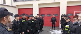 Zbiórka dwóch stojących na przeciwko siebie szeregów strażaków i pracowników cywilnych. W środku między szeregami stoi odchadzący na emeryturę st. ogn. Jacek Dolata. W lewym rogu bark Komendanta z pagonem. Wszyscy strażacy w czarnych mundurach dowódczo-sztabowych i koszarowych. Zbiórka odbywa się na placu Komendy z kostki brukowej, w tle czerwone bramy i żółta elewacja budynku Komendy.