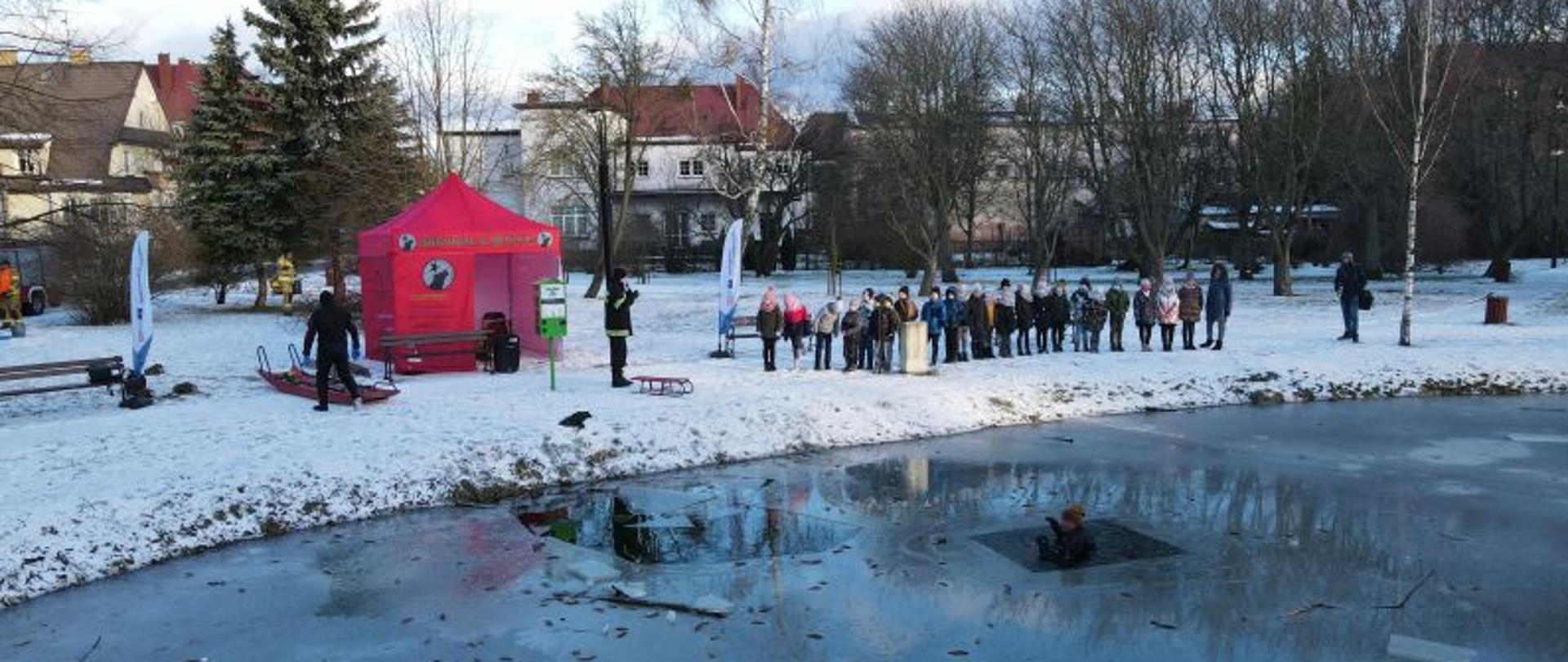 Zdjęcie wykonane w dzień z drona z niewielkiej wysokości. Widać na nim staw w parku miejskim na którego ośnieżonym brzegu stoją w szeregu dzieci z przedszkola. Tafla wody jest zamarznięta a w przeręblu jest zanurzony symulant. Na brzegu stoi czerwony namiot