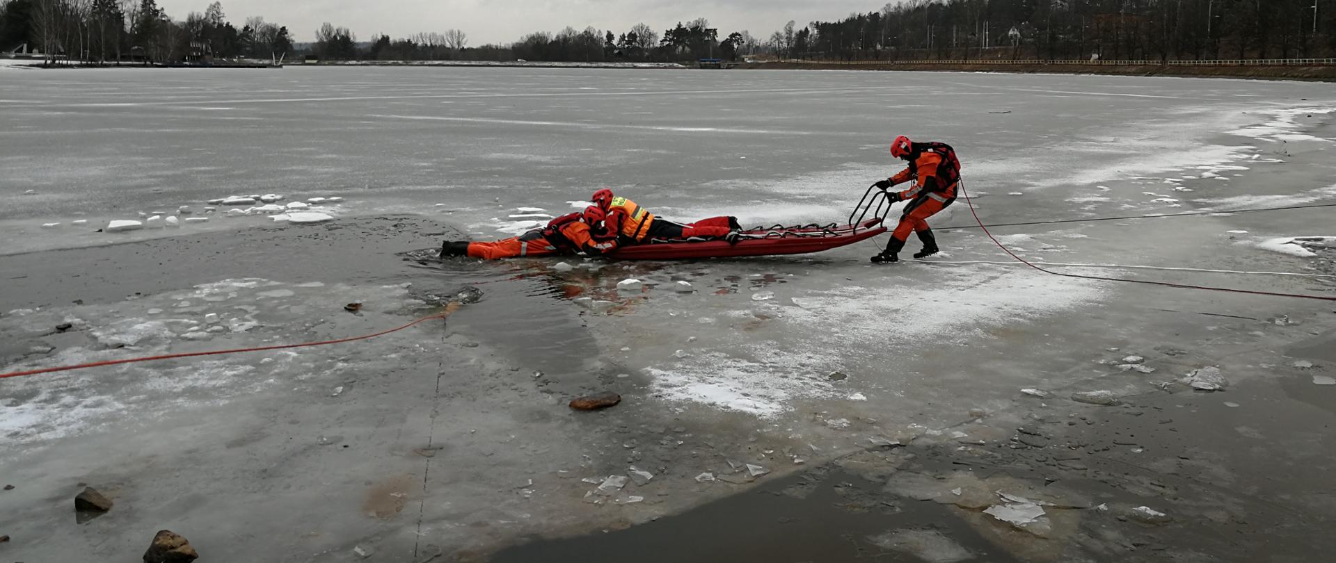 Na zdjęciu strażaków podczas ćwiczeń z ratownictwa lodowego. Dwóch strażaków wyciąga za pomocą sań lodowych poszkodowanego z wody. Wszyscy są zabezpieczeni linkami oraz ubrani w kamizelki asekuracyjne i specjalne ubrania do ratownictwa lodowego.