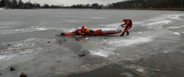 Na zdjęciu strażaków podczas ćwiczeń z ratownictwa lodowego. Dwóch strażaków wyciąga za pomocą sań lodowych poszkodowanego z wody. Wszyscy są zabezpieczeni linkami oraz ubrani w kamizelki asekuracyjne i specjalne ubrania do ratownictwa lodowego.