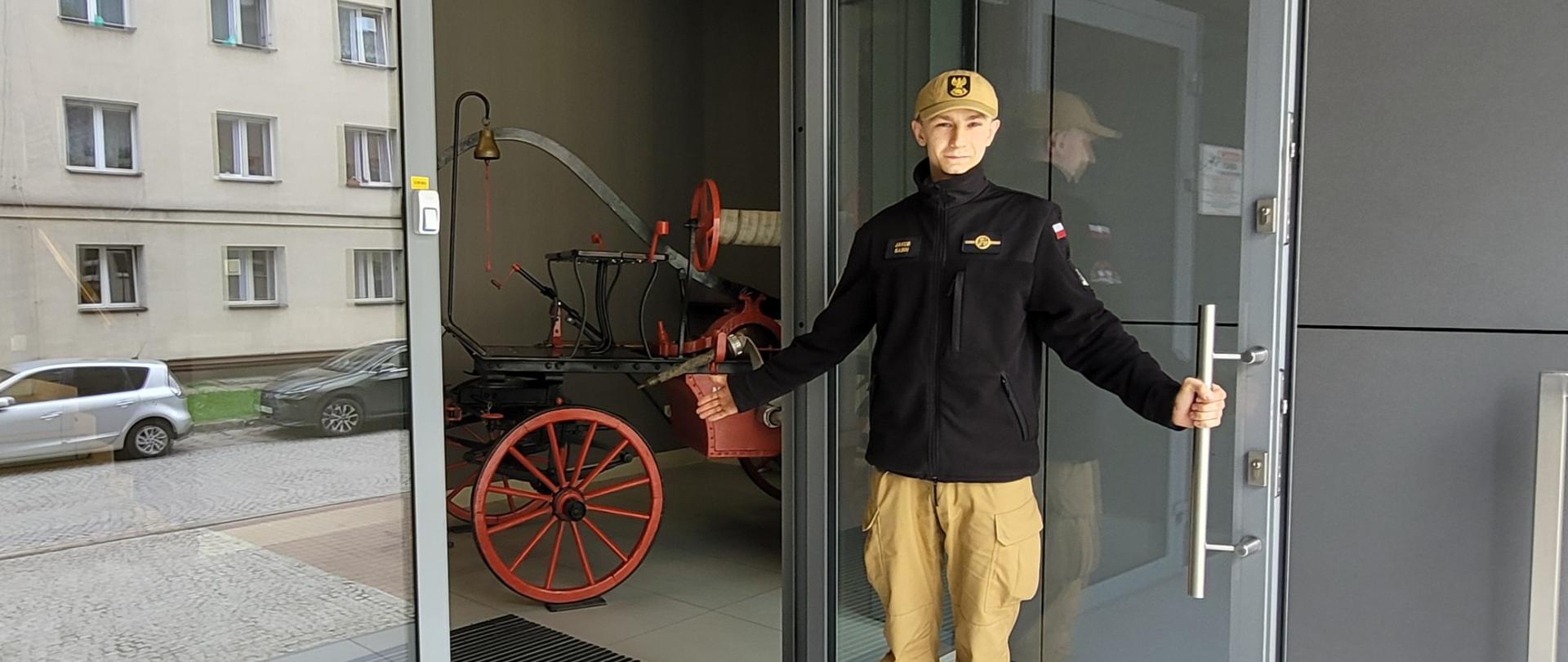 ubrany w umundurowanie słuchacz SA PSP w Krakowie otwiera drzwi wejściowe i serdecznym gestem zaprasza gości do środka