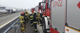 Grupa strażaków podczas instruktażu prowadzonego przez pracowników wykonawcy autostrady. W tle autostrada. Strażacy przy barierze energochłonnej.