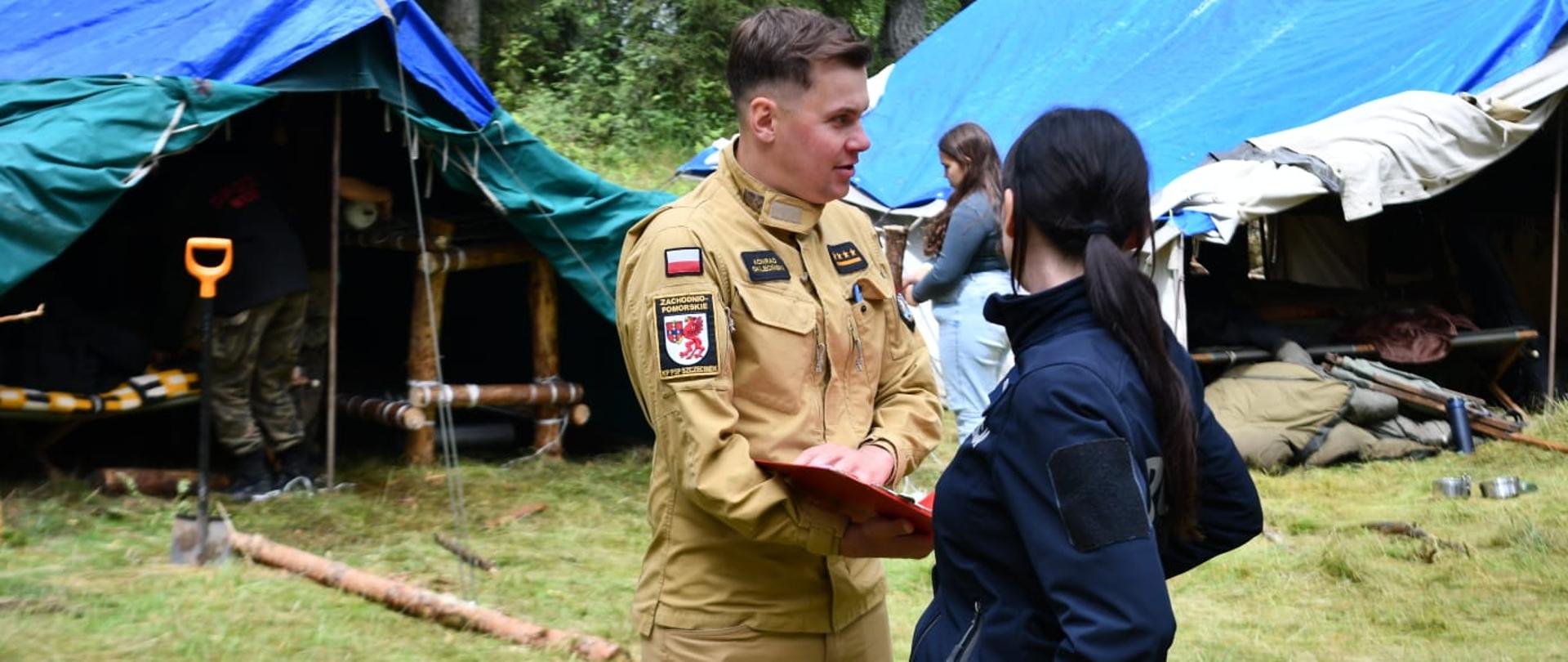 Strażak ubrany w mundur służbowy koloru piaskowego razem z Policjantką ubraną w mundur służbowy koloru granatowego stoją na tle namiotów. Sceneria odbywa się podczas kontroli obozu harcerskiego w lesie.