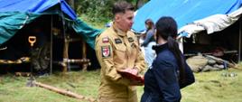 Strażak ubrany w mundur służbowy koloru piaskowego razem z Policjantką ubraną w mundur służbowy koloru granatowego stoją na tle namiotów. Sceneria odbywa się podczas kontroli obozu harcerskiego w lesie.