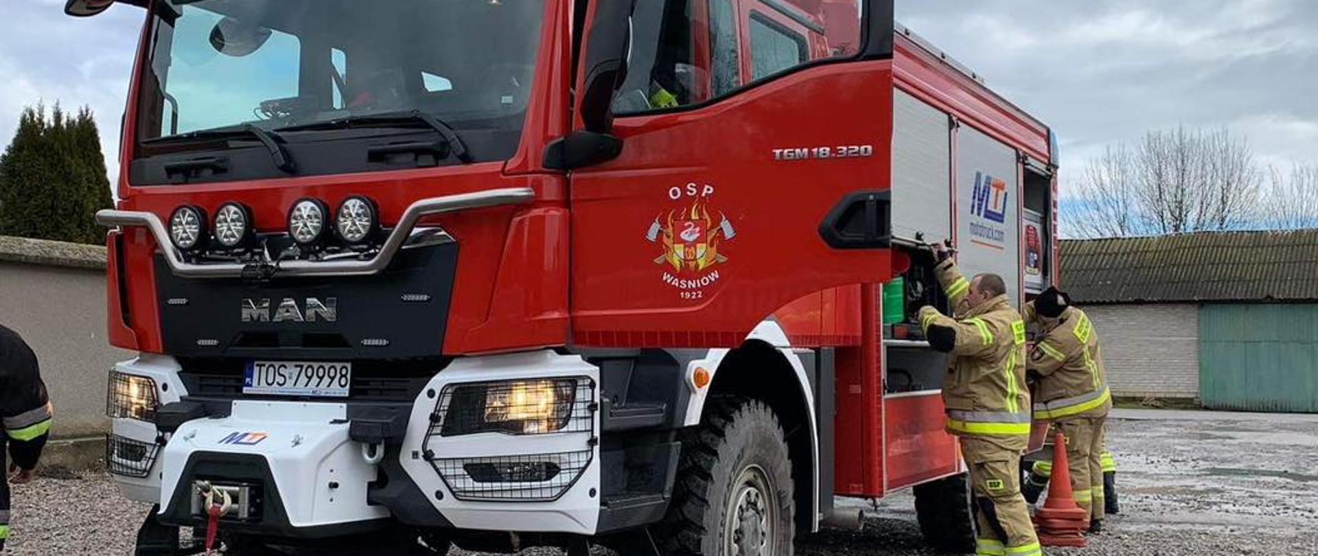 Na pierwszym planie widzimy samochód ratowniczo-gaśniczy OSP Waśniów, z którego skrytek dwóch ratowników wydostaje sprzęt. W tle widać murowany garaż a z boku betonowy płot