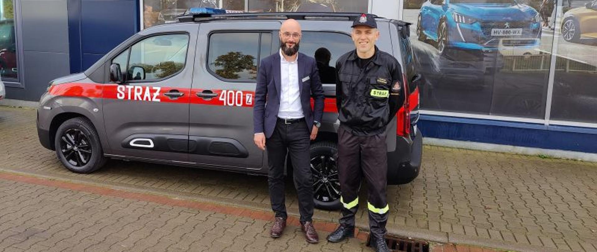 Zdjęcie wykonane na zewnątrz, przed budynkiem salonu samochodowego. Tło stanowi szara ściana. Jest jasno, słonecznie. Na pierwszym planie auto osobowe, minibus. Przed autem dwóch mężczyzn. Jeden ubrany w cywilne ubrania. Łysy z brodą, w okularach i marynarce. Obok po jego prawej stronie strażak w czarnym ubraniu w czapce. Obydwoje uśmiechnięci.
