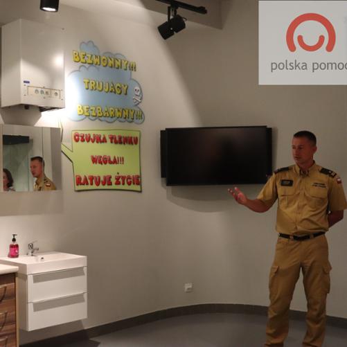 Strażak w mundurze prowadzi prezentację stojąc obok ekranu, plakatów informacyjnych i ekspozycji imitującej wyposażenie domu