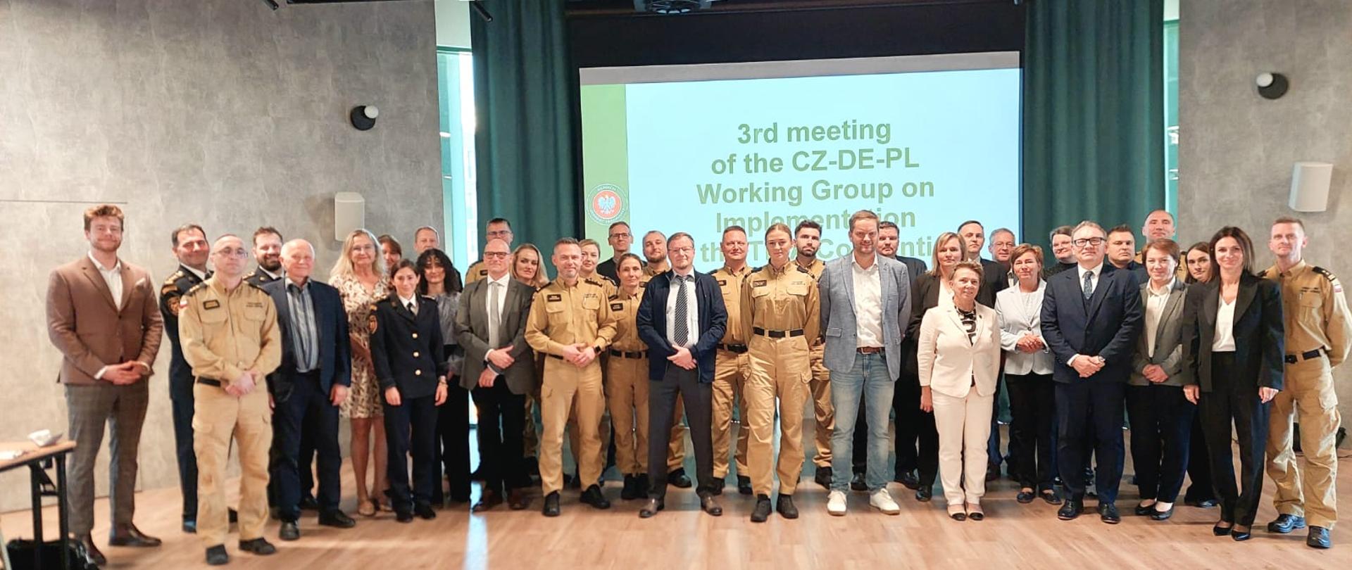 Grupa uczestników polsko-czesko-niemieckiego spotkania roboczego pozuje do wspólnego zdjęcia w sali konferencyjnej. Wśród uczestników są funkcjonariusze w mundurach PSP oraz przedstawiciele instytucji partnerskich. W tle ekran z informacją o trzecim spotkaniu grupy roboczej CZ-DE-PL.