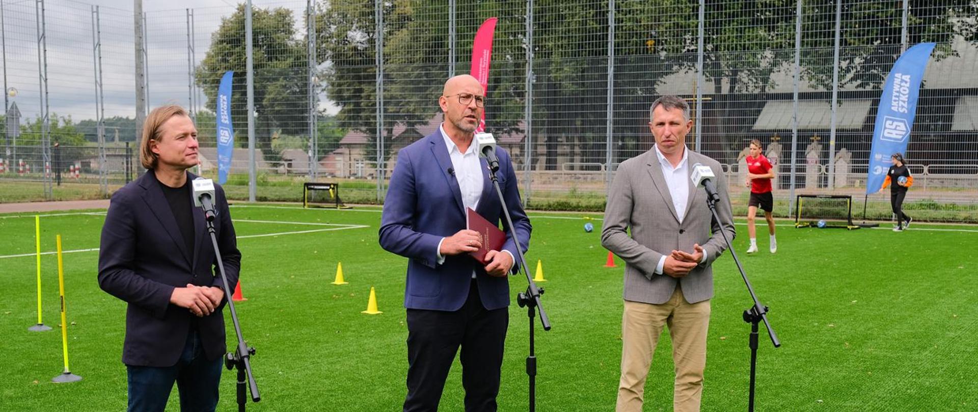 Three participants of the press conference (Minister of Sport and Tourism Jakub Rutnicki, Sławomir Szmal, President of the Polish Handball Federation and Mariusz Frankowski, Governor of Mazowieckie Voivodeshipstand) behind microphones on a football field.