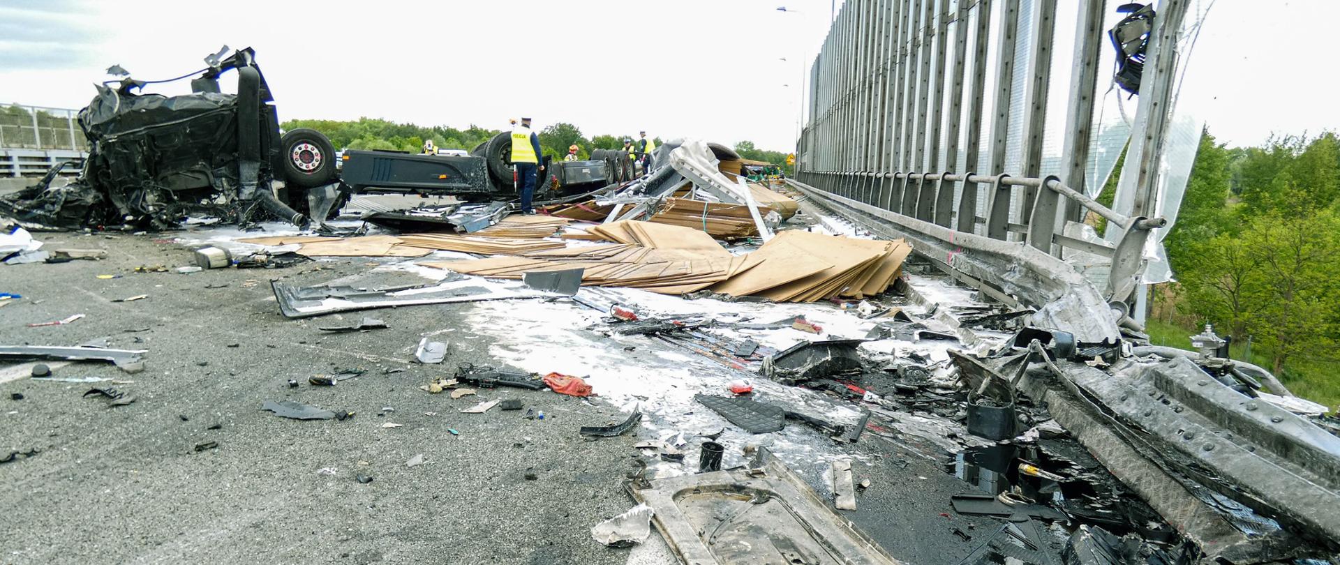 Na zdjęciu widoczny jest poważny wypadek drogowy na moście. Kilka pojazdów jest przewróconych i poważnie uszkodzonych. Na jezdni rozrzucone są fragmenty wraków i inne szczątki. Barierki mostu są wygięte lub połamane. W tle znajdują się służby ratunkowe, które oceniają sytuację. Scena przedstawia chaos i zniszczenia po zdarzeniu drogowym.
