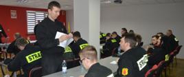 Egzamin końcowy. Świetlica Komendy Powiatowej PSP w Rawiczu. Przy stołach siedzą egzaminowani strażacy. Przewodniczący komisji egzaminacyjnej rozdaje im testy wraz z kartami odpowiedzi. 