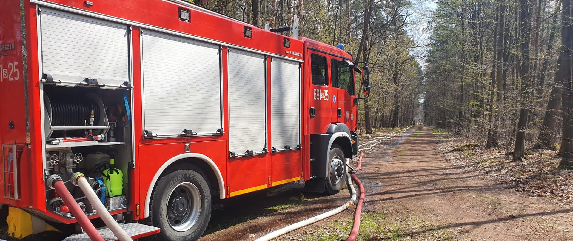 Samochód strażacki stoi na leśnej drodze, od niego rozwinięte węże wzdłuż drogi. W głębi przy wężach dwóch strażaków.
