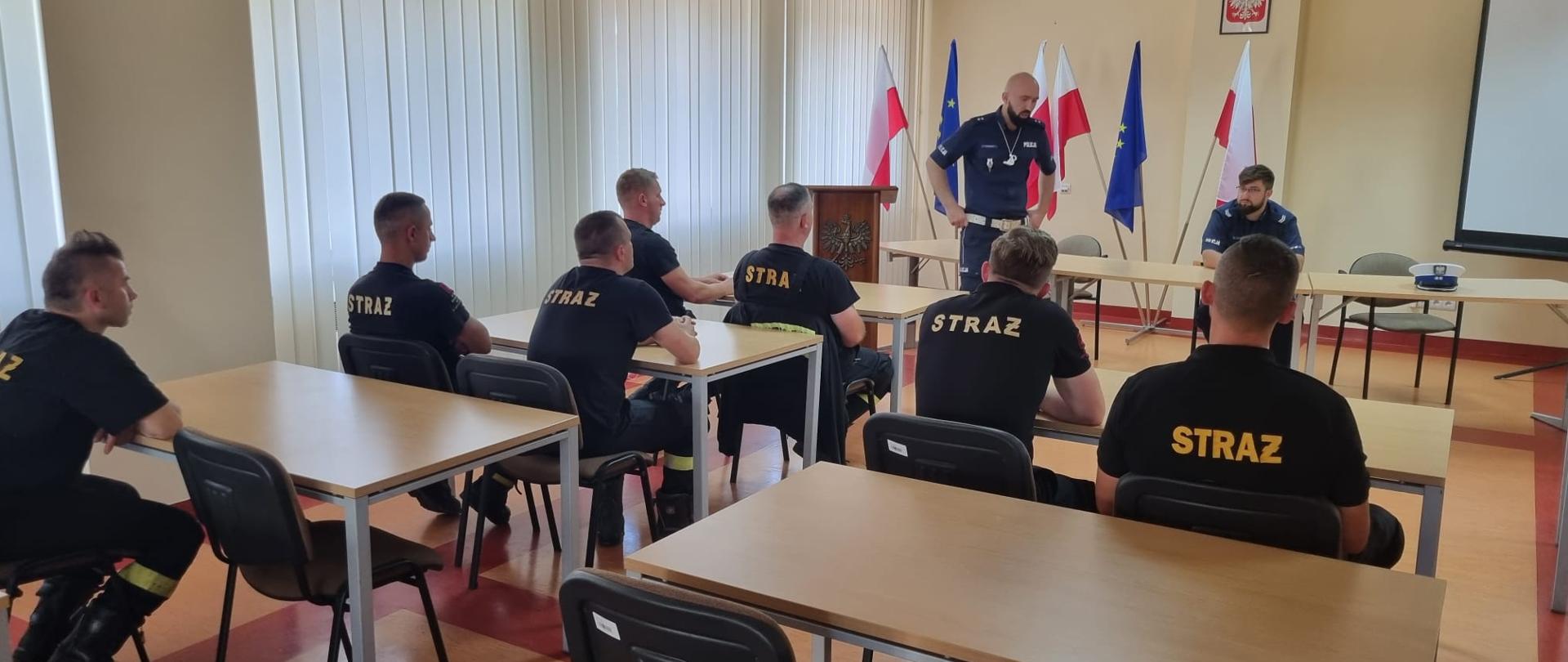 Zdjęcie przedstawia stojącego funkcjonariusza z Wydziału Ruchu Drogowego Komendy Powiatowej Policji w Jędrzejowie, który prowadzili prelekcje z zakresu zasad bhp i przepisów dotyczących bezpieczeństwa ruchu drogowego. Na świetlicy przy stolikach siedzą strażacy. Koło nich przy stoliku siedzi kolejny funkcjonariusz policji.