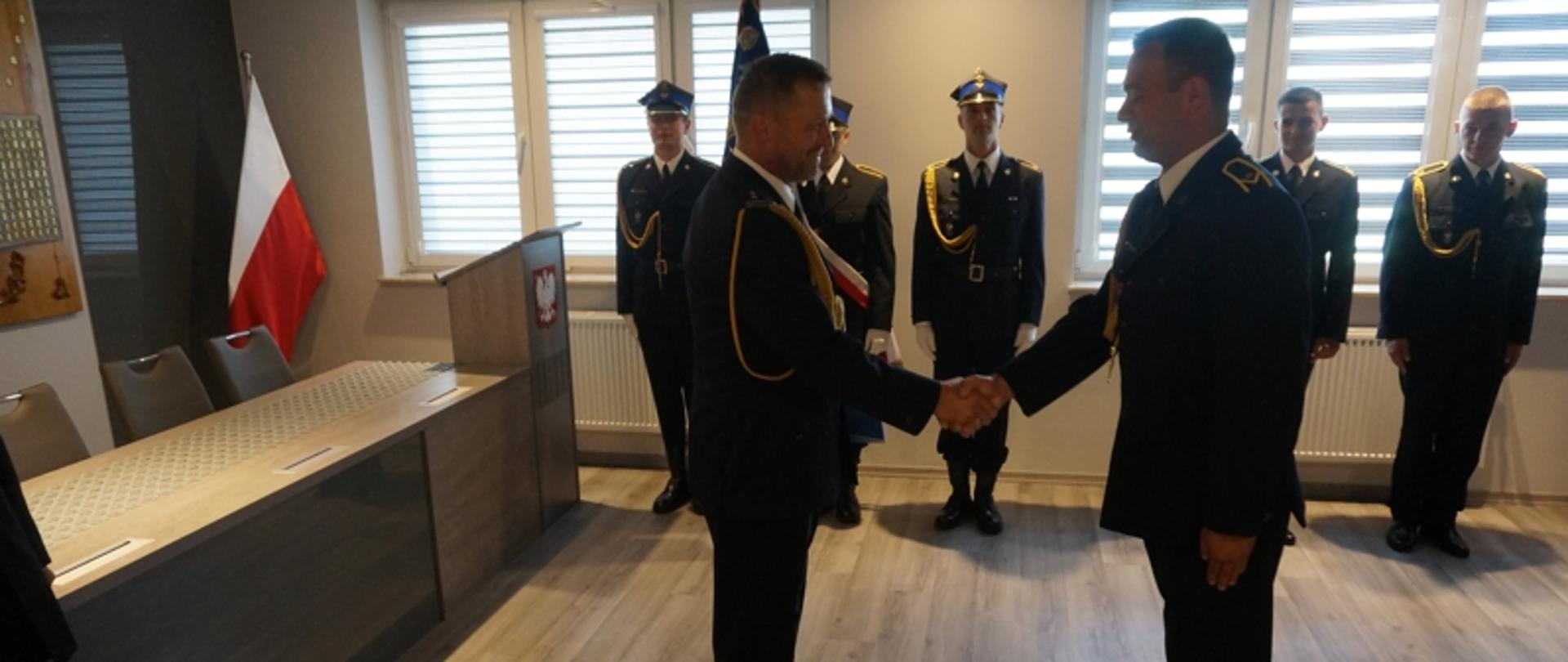 Komendant Powiatowy PSP bryg. Robert Lebioda wręcza akt nadania pierwszego stopnia w korpusie aspirantów PSP st. ogn. Damianowi Wyce podczas uroczystej zbiórki. Uczestnicy ubrani w umundurowanie wyjściowe. W tle poczet sztandarowy Komendy Powiatowej PSP w Stalowej Woli.