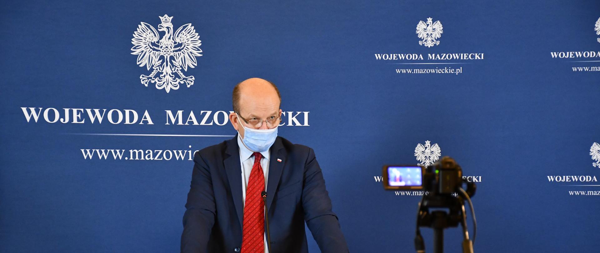 Na zdjęciu znajduje się mężczyzna, ubrany w granatowy garnitur, jasną koszulę i czerwony krawat. Mężczyzna ten stoi przed kamerą, będąc jednocześnie za mównicą (na mównicy znajduje się godło Polski oraz napis "Wojewoda Mazowiecki"). W tle ścianka wizerunkowa w kolorze jasnogranatowym. Na ściance znajdują się logotypy Wojewody, czyli godło Polski i napis "Wojewoda Mazowiecki"