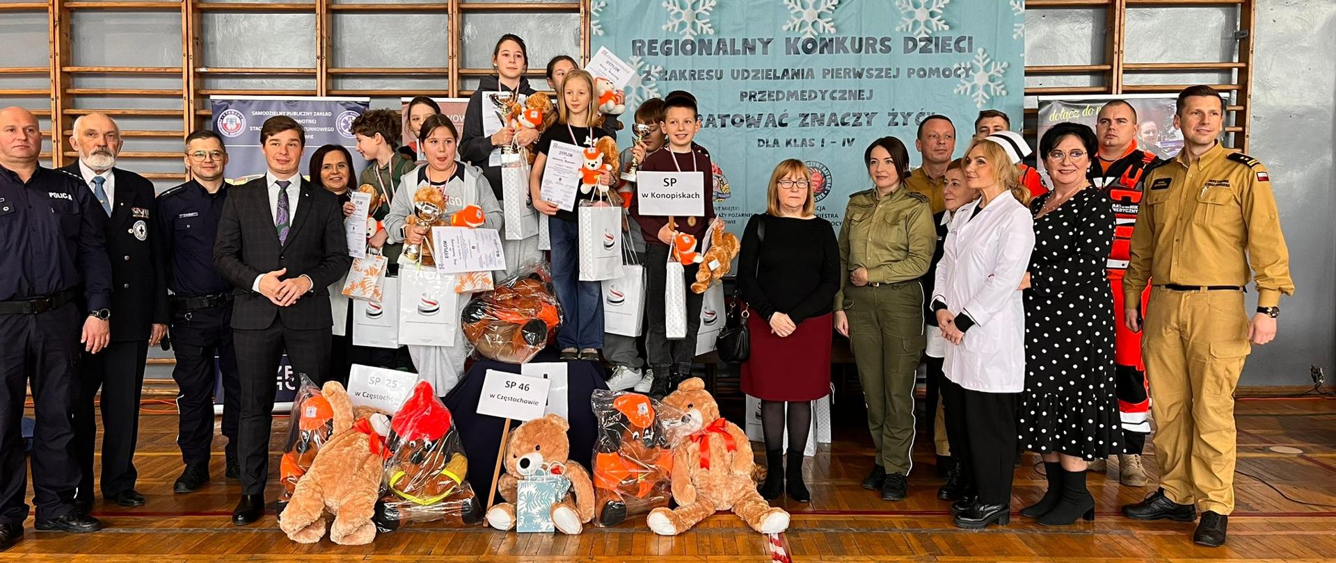Na zdjęciu widać grupowe, pamiątkowe zdjęcie wykonane w sali gimnastycznej. W centrum stoją dzieci – laureaci regionalnego konkursu z zakresu udzielania pierwszej pomocy przedmedycznej. Trzymają dyplomy, puchary i torby z nagrodami, a przed nimi na podłodze leżą duże pluszowe misie ubrane w elementy strażackie.
Po obu stronach dzieci stoją dorośli opiekunowie i organizatorzy: strażacy w mundurach Państwowej Straży Pożarnej, policjant, ratownicy medyczni oraz przedstawiciele instytucji w strojach służbowych i cywilnych. W tle widoczny jest baner z napisem informującym o regionalnym konkursie pierwszej pomocy oraz drabinki gimnastyczne.