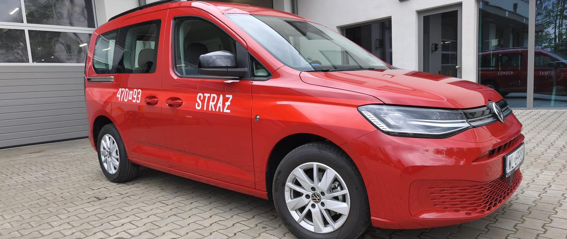 Zdjęcie przedstawia samochód osobowy w kolorze czerwonym. Widoczny jest prawy bok oraz częściowo przód pojazdu. Na drzwiach widoczna naklejka z napisem Straż oraz numery 470m93. W tle widać bramy garażowe budynku. Pojazd stoi na kostce brukowej