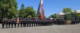 Zdjęcie przedstawia uroczysty apel strażaków na placu Centralnej Szkoły Państwowej Straży Pożarnej. Na pierwszym planie widać kilkudziesięciu strażaków ustawionych w równych szeregach. Większość z nich ubrana jest w galowe, czarne mundury z czapkami i złotymi sznurami na ramieniu. Po lewej stronie stoi strażak trzymający sztandar. Po prawej stronie grupy widoczna jest także sekcja strażaków w piaskowych mundurach służbowych. W tle stoją czerwone pojazdy strażackie, a za nimi samochód podnośnik z zawieszonym banerem z napisem: „Centralna Szkoła Państwowej Straży Pożarnej” i logo PSP. Na jej szczycie umieszczony jest kosz z podnośnika strażackiego, który unosi się wysoko w górę. Dookoła placu rosną drzewa, a po prawej stronie znajduje się budynek szkoły. Pogoda jest słoneczna, niebo bezchmurne, co podkreśla podniosły charakter wydarzenia.