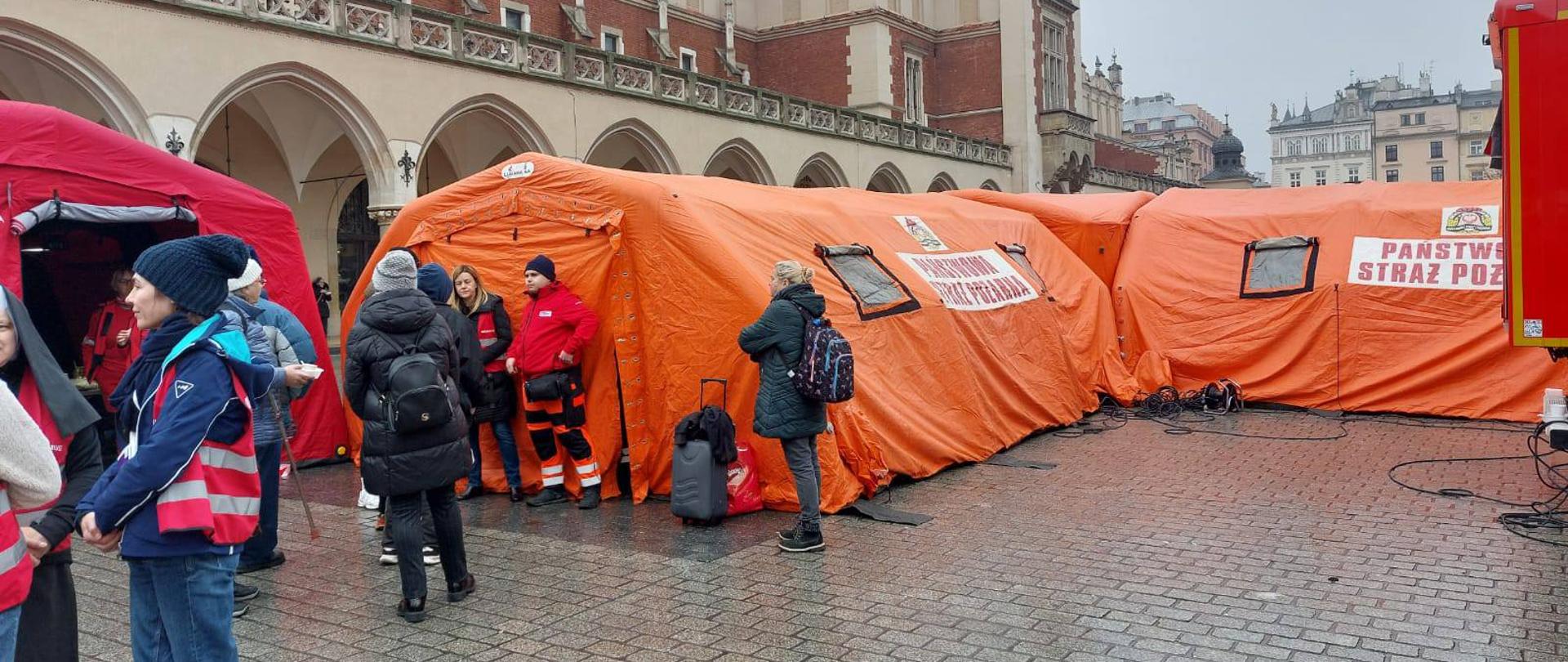 Zimowy poranek na krakowskim Rynku. Na pierwszym planie duże, czerwone namioty strażackie (Szpital Polowy). Wokół nich ludzie w ubraniach cywilnych zimowych oraz wolontariusze w czerwonych kamizelkach W tle widać oświetlone Sukiennice i tłum ludzi.