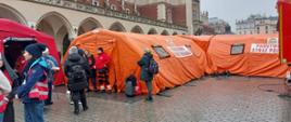 1 Zimowy poranek na krakowskim Rynku. Na pierwszym planie duże, czerwone namioty strażackie (Szpital Polowy). Wokół nich ludzie w ubraniach cywilnych zimowych oraz wolontariusze w czerwonych kamizelkach W tle widać oświetlone Sukiennice i tłum ludzi.