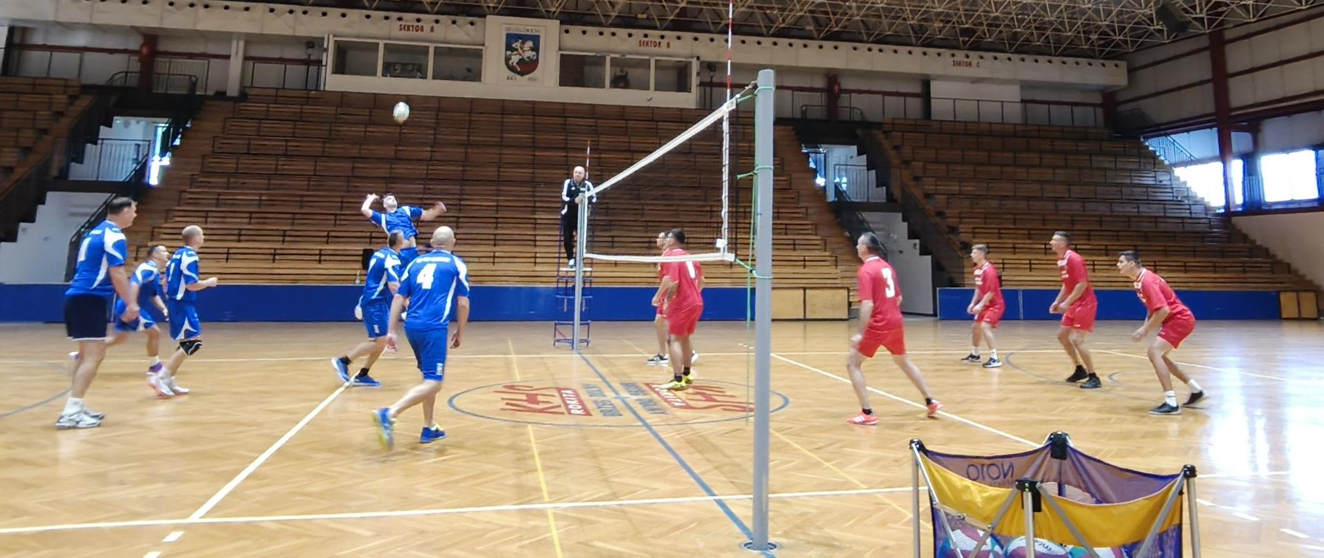 Zdjęcie przedstawia mecz siatkówki mężczyzn rozgrywany w dużej hali sportowej. Zawodnicy po obu stronach siatki ubrani są w niebieskie stroje sportowe. Po lewej stronie zawodnik odbija piłkę w stronę siatki, podczas gdy inni gracze obserwują akcję. W tle widoczne są puste, drewniane trybuny oraz sędzia stojący przy siatce na podwyższeniu.