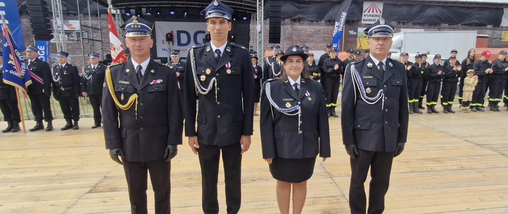 Na zdjęciu, zrobionym w plenerze, widać cztery osoby ubrane w odświętne, wyjściowe mundury strażackie. Stoją one na drewnianym podeście przed dużą sceną, na której w tle widać grupę innych strażaków druhów ze sztandarami. Pierwszy od lewej stoi śląski komendant wojewódzki państwowej straży pożarnej. Po prawej stoją druhowie w wyjściowych czarnych mundurach wśród nich jedna kobieta druhna. Wszyscy pozują do zdjęcia w postawie na baczność.