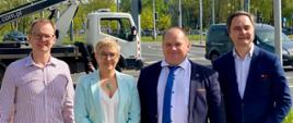 Wiceminister Mrówczyńska i trzech mężczyzn stoją na chodniku, w tle widać sygnalizację świetlną i zielone drzewa.