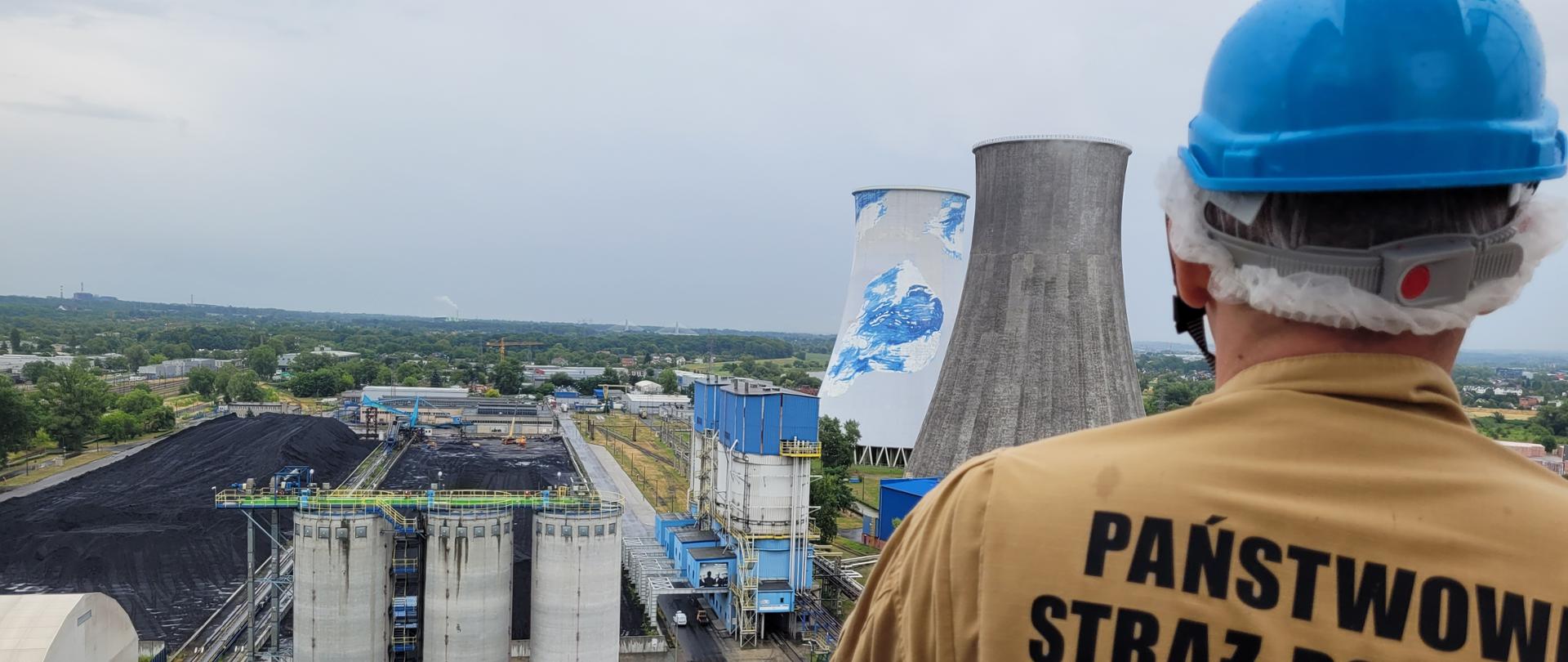 Stojący plecami do fotografa strażak Państwowej Straży Pożarnej z niebieskim kaskiem na głowie ogląda z podwyższenia infrastrukturę zakładu przemysłowego: kominy, hale, elementy technologiczne, składowisko (hałda) węgla. 