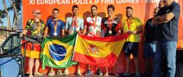 Ceremonia dekoracji medalowej na podium zawodów XI European Police & Fire Games w Vigo. Sześciu zawodników ze srebrnymi i brązowymi medalami na szyi pozuje na scenie. Trzymają przed sobą rozpostarte flagi Niemiec, Brazylii oraz Hiszpanii. W tle znajduje się pomarańczowa ścianka z nazwą wydarzenia, a obok podium stoją organizatorzy.