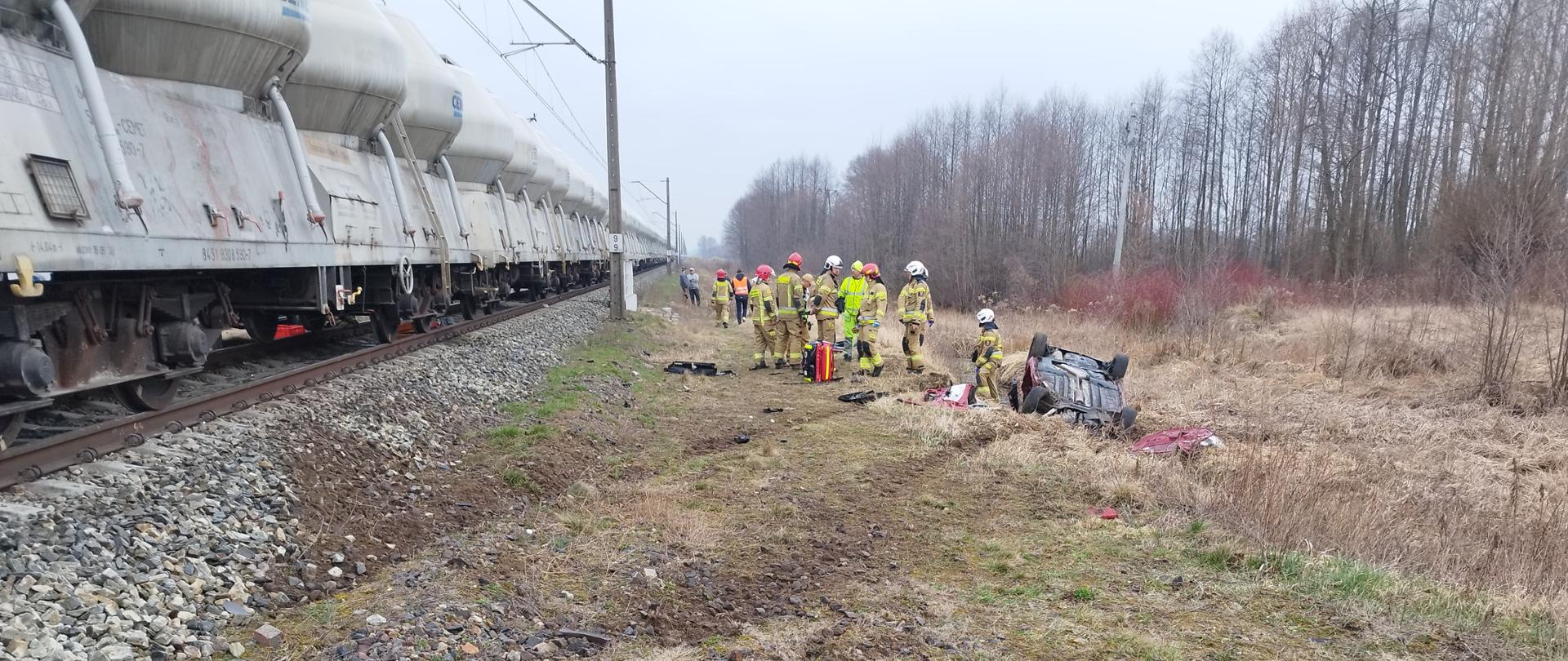 Zdjęcie przedstawia strażaków stojących pomiędzy wagonami pociągu towarowego a samochodem na dachu. Auto znajduje sie w rowie. Strażacy ubrani są w ubrania specjalne.