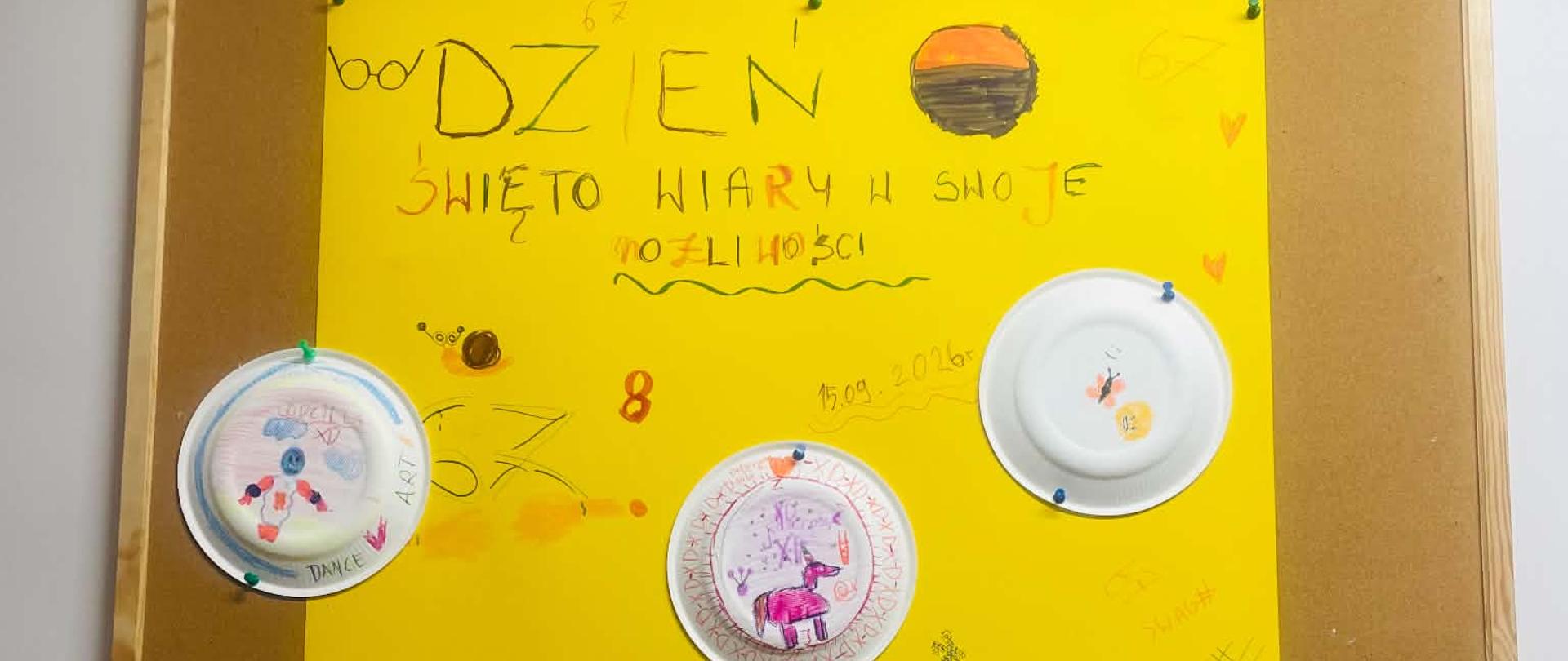 Zdjęcie plakatu. Na żółtej kartce formatu A3 zamieszczono napis: "Dzień święto wiary w swoje możliwości" oraz 9 białych papierowych talerzyków na których dziewczynki namalowały swoje rysunki.