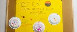 Zdjęcie plakatu. Na żółtej kartce formatu A3 zamieszczono napis: "Dzień święto wiary w swoje możliwości" oraz 9 białych papierowych talerzyków na których dziewczynki namalowały swoje rysunki.