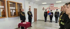 Na zdjęciu w mundurach galowych po lewej komendant powiatowy wraz z zastępcą po środku poczet sztandarowy ze sztandarem. Po prawej strażacy ubrani w czarne ubrania koszarowe uczestniczący w uroczystość ślubowania na sali konferencyjnej.