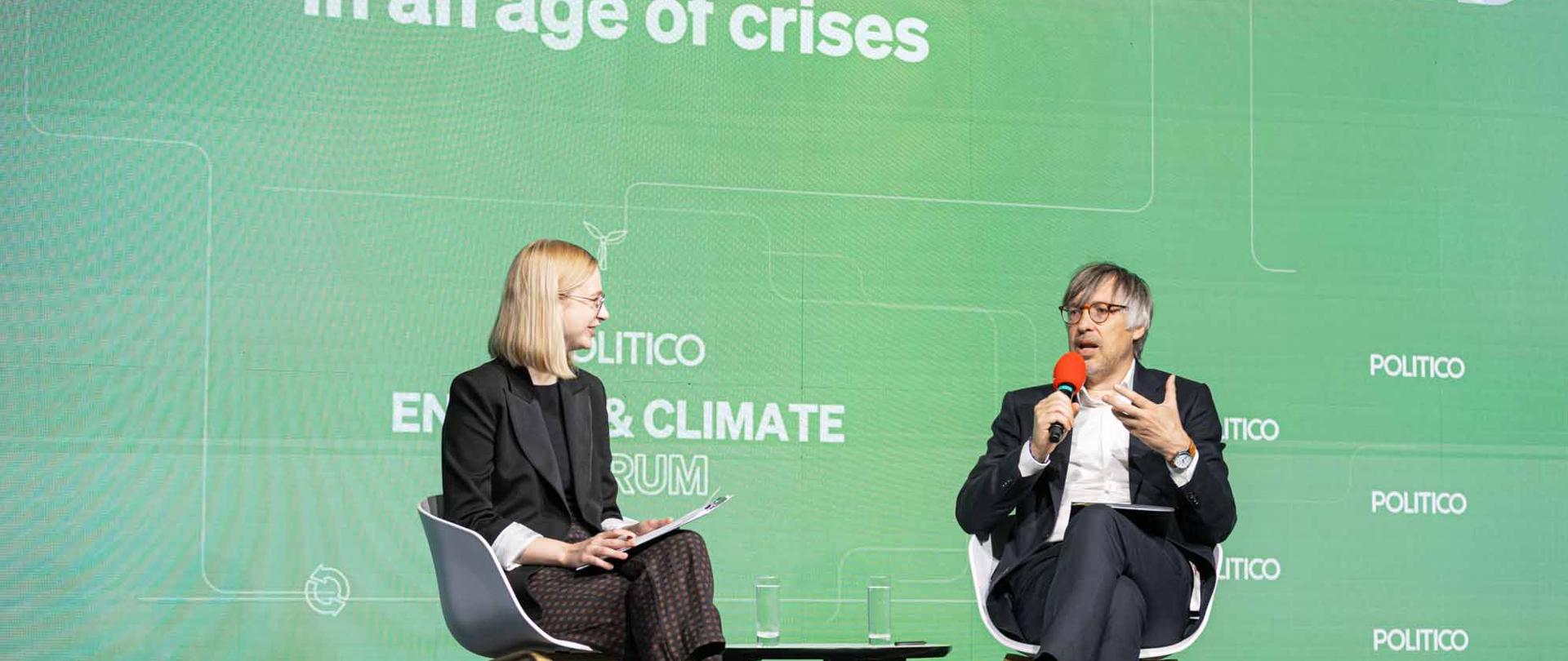 Wiceminister klimatu i środowiska Krzysztof Bolesta podczas POLITICO Energy & Climate Forum. Zdjęcie autorstwa POLITICO Europe. Na zdjęciu wiceminister rozmawia z dziennikarką i żywo gestykuluje. Za nim znajduje się ekran z napisem ,,Delivering climate action in an age of crises".