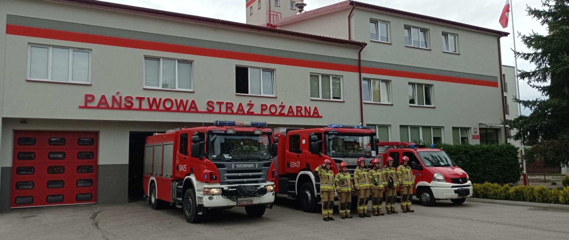 Na zdjęciu widoczny budynek Komendy Powiatowej PSP w Sochaczewie. Przed garaże wyjechały trzy samochody pożarnicze, przed którymi w szeregu stoją strażacy w ubraniach specjalnych i hełmach. Dowódca oddaje honor.