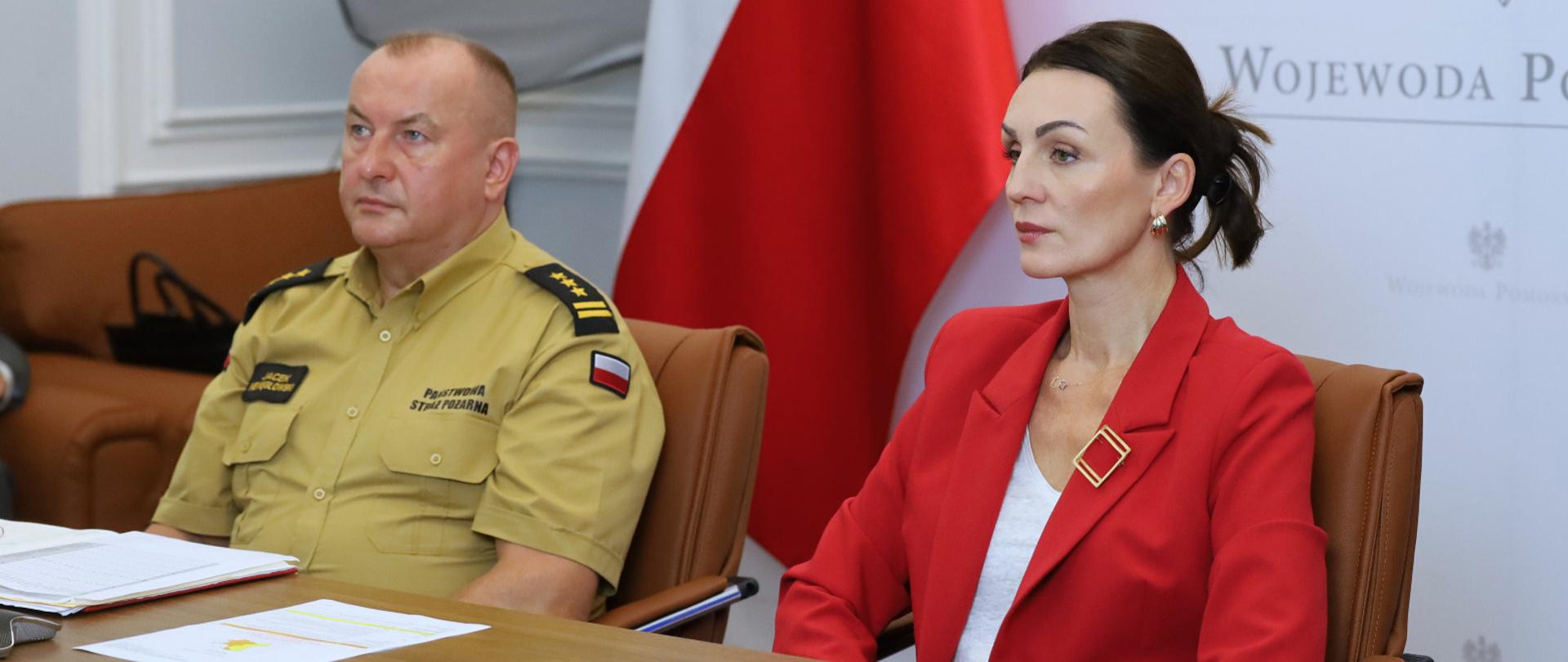 kobieta w czerwonej marynatce i mężczyzna w mundurze strażaka patrzą na ekran telewizora