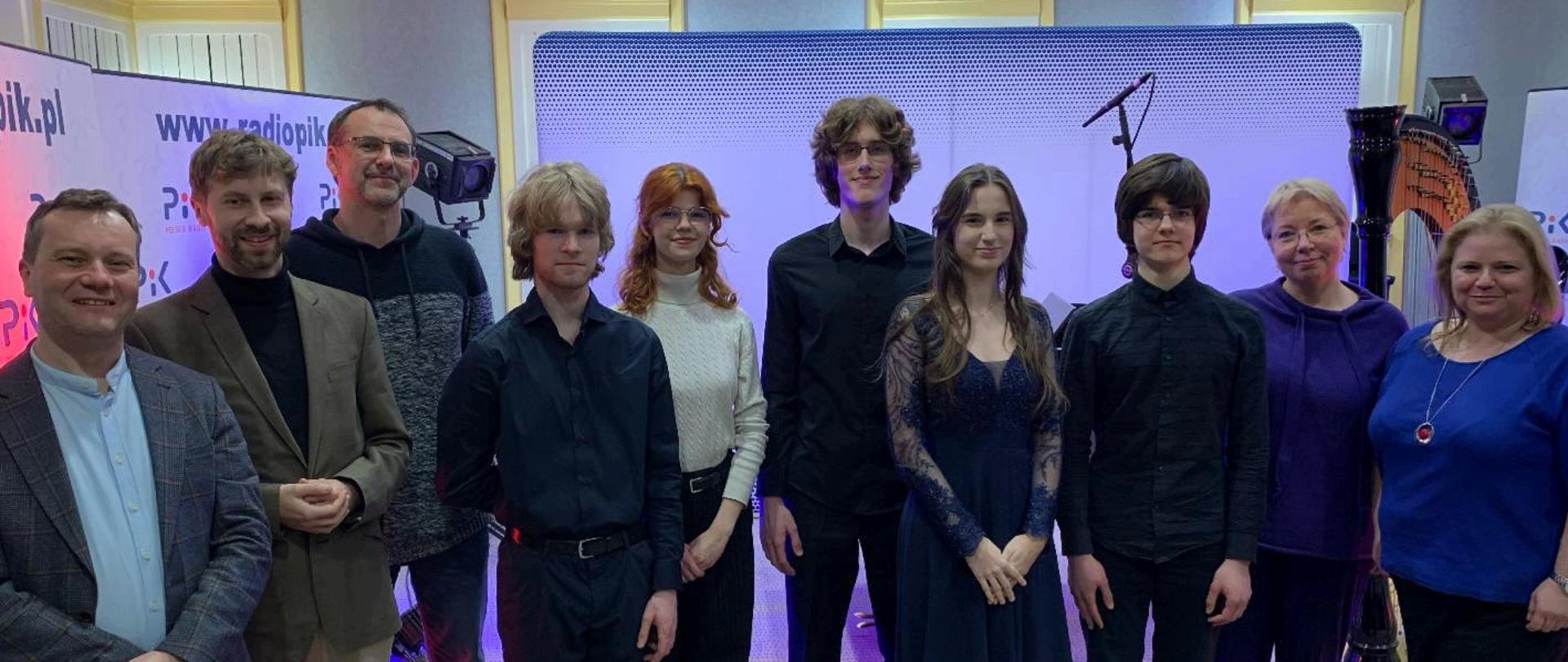 na zdjęciu jest półmrok otoczony różowym i niebieskim światłem na pierwszym planie stoi cała grupa osób w która brała udział w wywiadzie w radiu