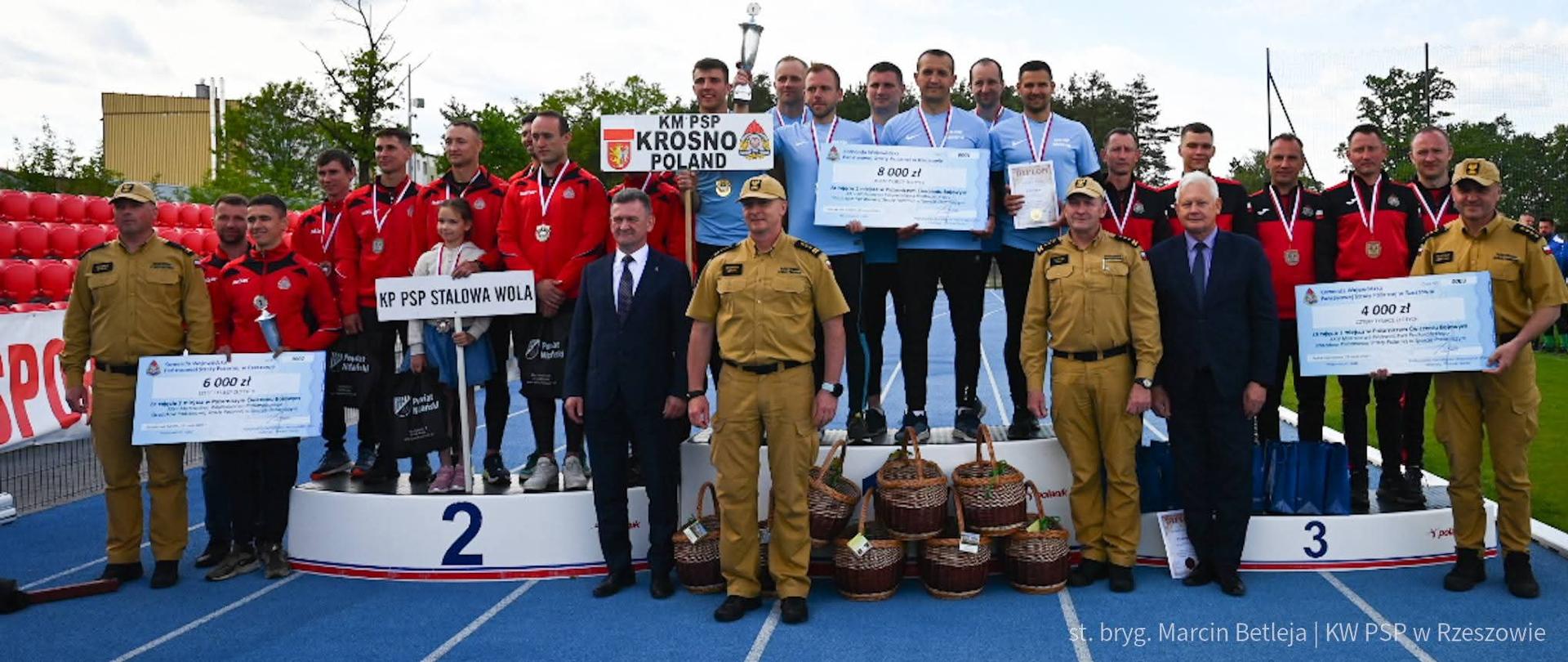 Kolorowa fotografia wykonana w słoneczny dzień na bieżni stadionu. Przedstawia najlepsze drużyny województwa podkarpackiego strażaków PSP w sporcie pożarniczym. Trzy najlepsze drużyny w dresach stoją na podium oraz strażacy w piaskowych ubraniach i osoby cywilne wręczający nagrody dyplomy oraz czeki pieniężne.