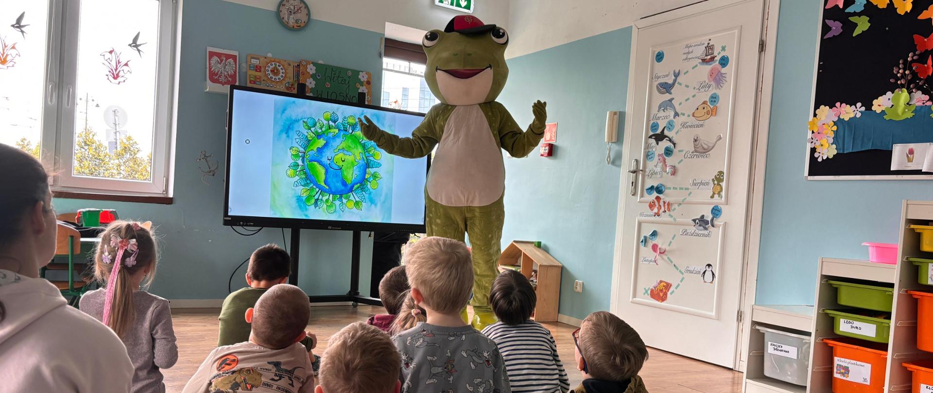 Żywa maskotka Żabka, stoi obok monitora z wyświetloną grafiką Dnia Ziemi, przed nią siedzą na macie dzieci i słuchają.