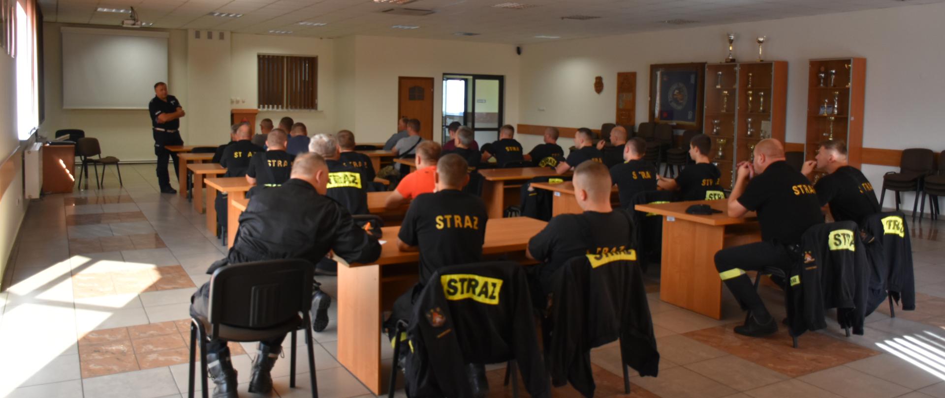 Zdjęcie obrazuje strażaków powiatu skarżyskiego uczestniczących w szkoleniu z zakresu prowadzenia pojazdów uprzywilejowanych. Prowadzącym zajęcia jest policjant. Strażacy siedzą w auli słuchając szkolenia. 