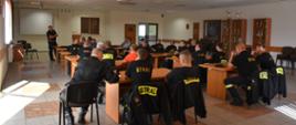 Zdjęcie obrazuje strażaków powiatu skarżyskiego uczestniczących w szkoleniu z zakresu prowadzenia pojazdów uprzywilejowanych. Prowadzącym zajęcia jest policjant. Strażacy siedzą w auli słuchając szkolenia. 