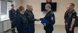 Zdjęcie przedstawia moment wręczania awansów służbowych funkcjonariuszom PSP. Komendant Powiatowy gratulując wyróżnienia ściska jednemu z nich dłoń. Wszyscy ubrani są w czarne stroje służbowe. Po lewej stronie widać fragment białego grzejnika oraz przysłonięte roletami okno. W tle widnieje biała ściana, na której zawieszony jest telewizor a nad nim godło Polski.