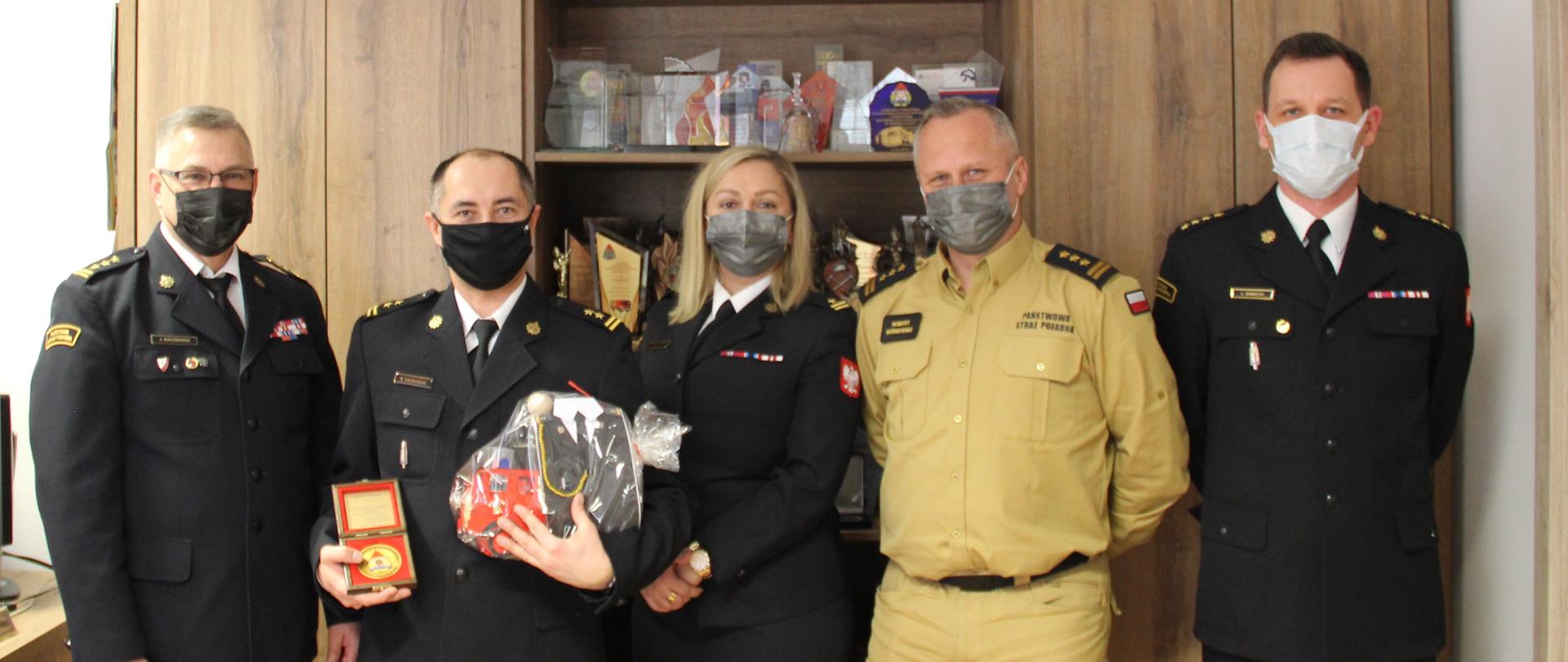 Zdjęcie wykonano w gabinecie Komendanta Wojewódzkiego PSP w Toruniu. Na zdjęciu w szeregu stoją od prawej Kujawsko-Pomorski Komendant Wojewódzki PSP st. bryg. Jacek Kaczmarek, bryg. Michał Gołębiowski (trzyma w rękach upominki oraz medal okolicznościowy), mł. bryg. Ewa Raciniewska Naczelnik Wydziału Kadr KW PSP w Toruniu, Zastępca Kujawsko-Pomorskiego Komendanta Wojewódzkiego PSP st. bryg. Robert Wiśniewski oraz Kierownik Sekcji SK KW PSP w Toruniu st. kpt. Leszek Baniecki.