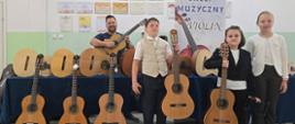 Zdjęcie grupowe w sali lekcyjnej lub koncertowej. Trójka dzieci – chłopak w kamizelce i dwie dziewczynki w eleganckich strojach – stoi z przodu, trzymając w rękach gitary klasyczne. Za nimi, po lewej stronie, stoi uśmiechnięty, brodaty mężczyzna, również trzymający gitarę. W tle znajduje się stół nakryty ciemnoniebieskim obrusem, na którym wyeksponowano kolejne gitary. Na ścianie wiszą liczne dyplomy w ramkach oraz baner reklamowy z napisem „SKLEP MUZYCZNY VIOLIN” przedstawiający gryf skrzypiec.