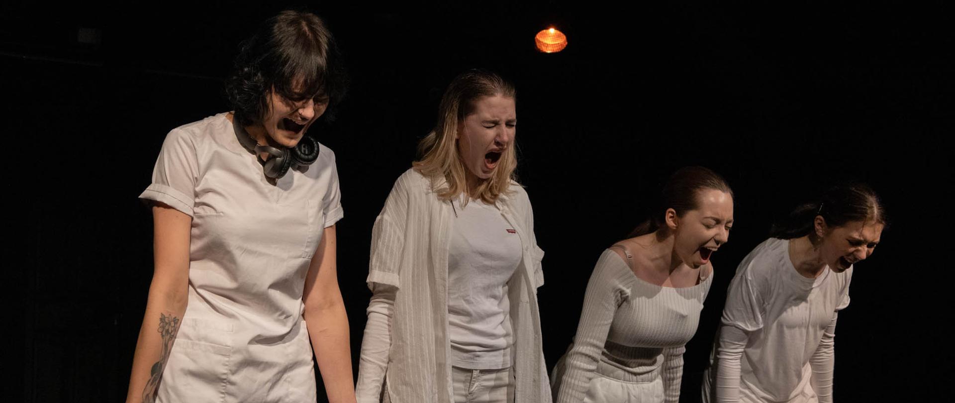 Zdjęcie prezentuje 4 aktorki w ciemnej sali teatralnej. Aktorki są zastane na zdjęciu w pozycji stojącej. Krzyczą. 